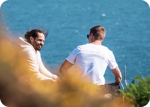 Two men sitting down and talking while surrounded by shrubbery with the sea in the background