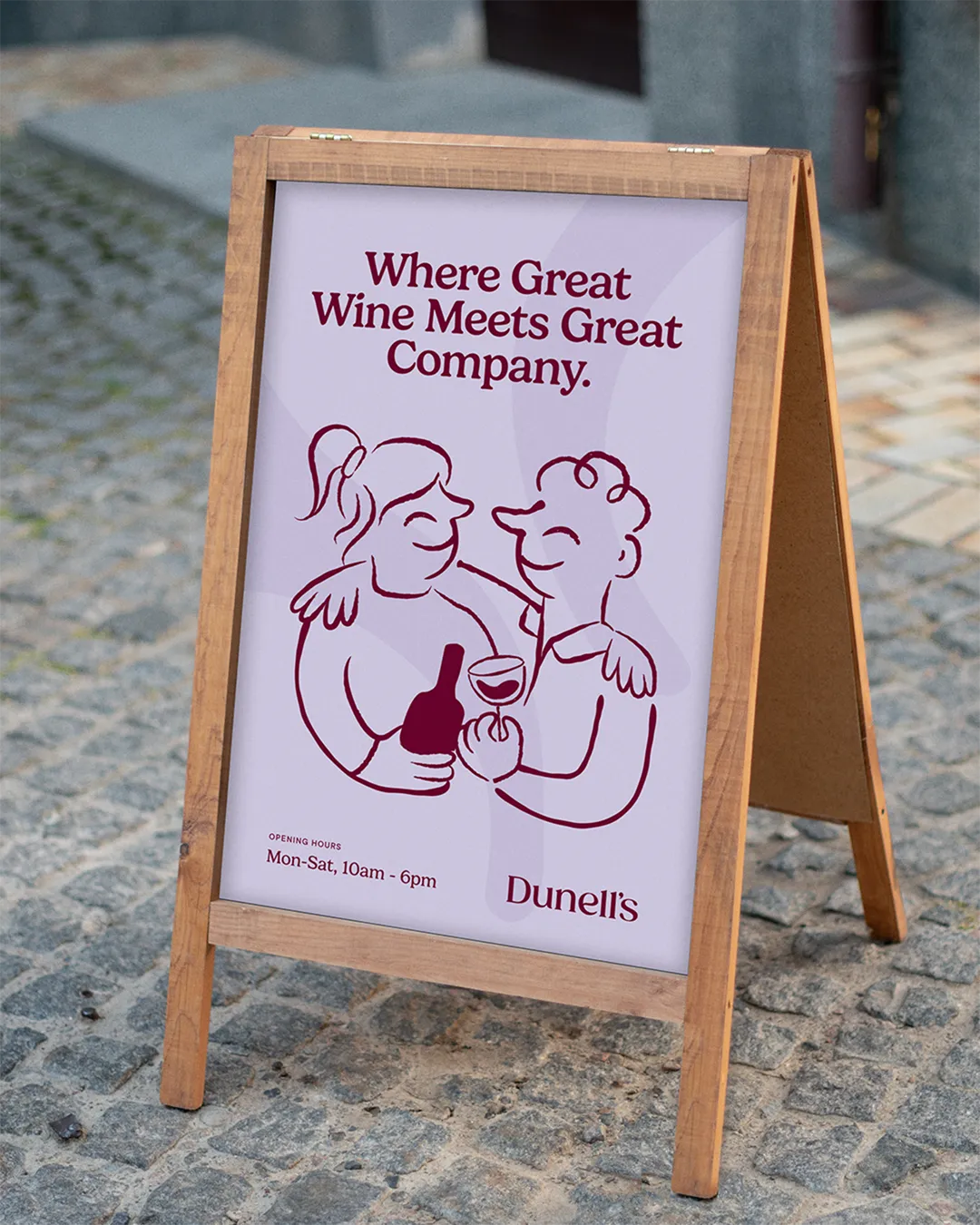 Wooden A-frame sidewalk sign with an illustration of two people holding a wine bottle and glass, text reading 'Where Great Wine Meets Great Company' and business hours 'Mon-Sat, 10am - 6pm'.