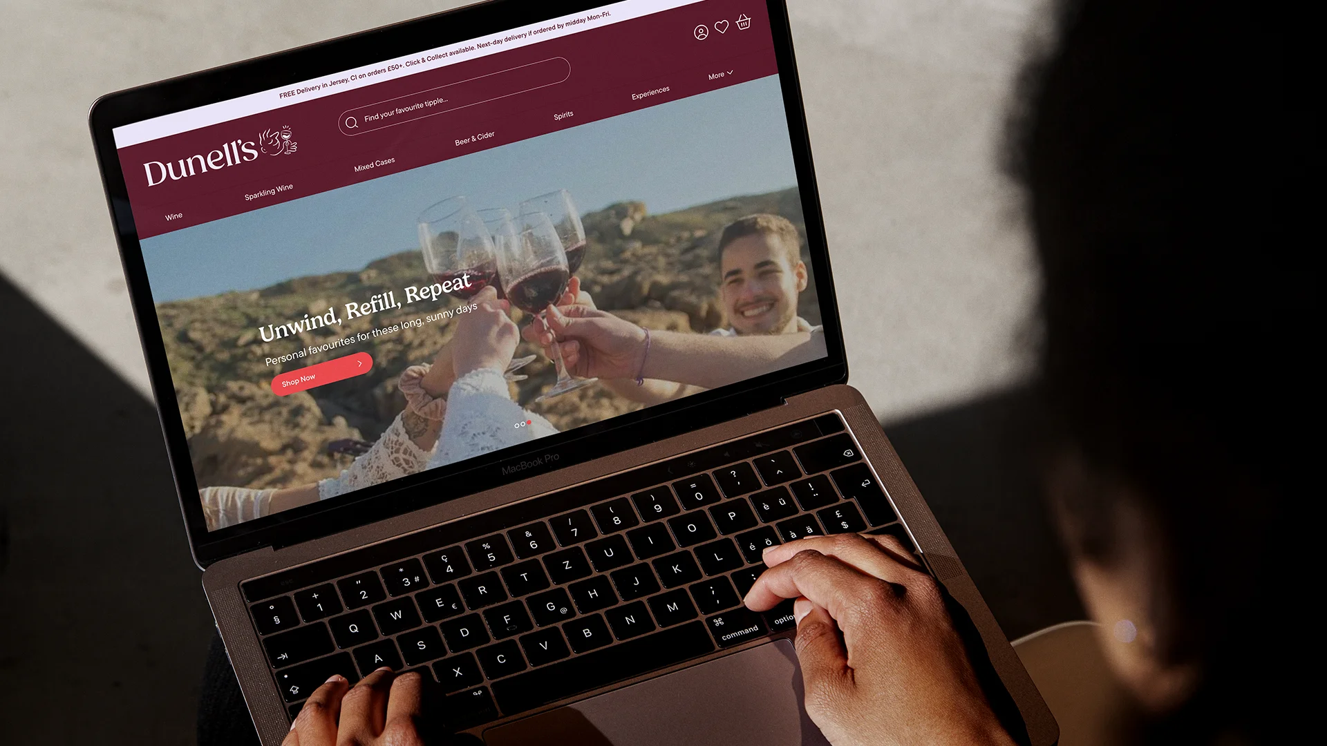 Person typing on a MacBook Pro with Dunell's website displaying a toast with wine glasses on screen.
