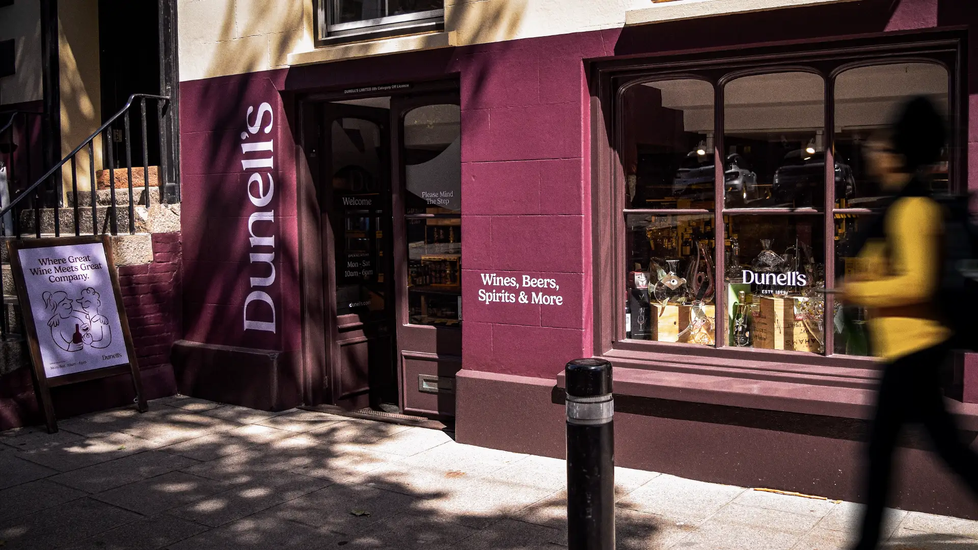 Exterior of Dunell's wine and spirits shop with signage and a blurred pedestrian walking by.
