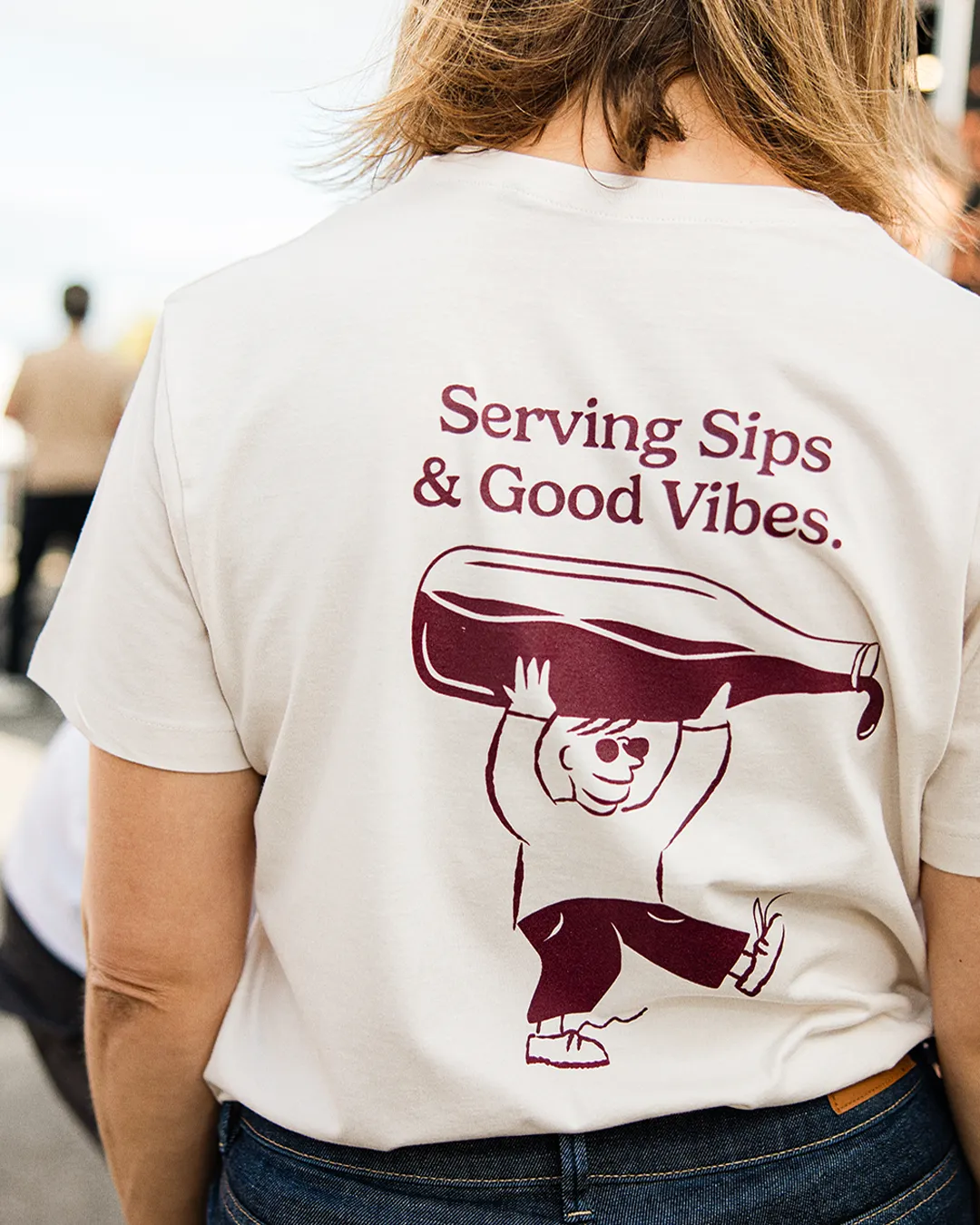 Back view of a person wearing a white T-shirt with a graphic of a cartoon figure carrying a large wine bottle and the text 'Serving Sips & Good Vibes.'