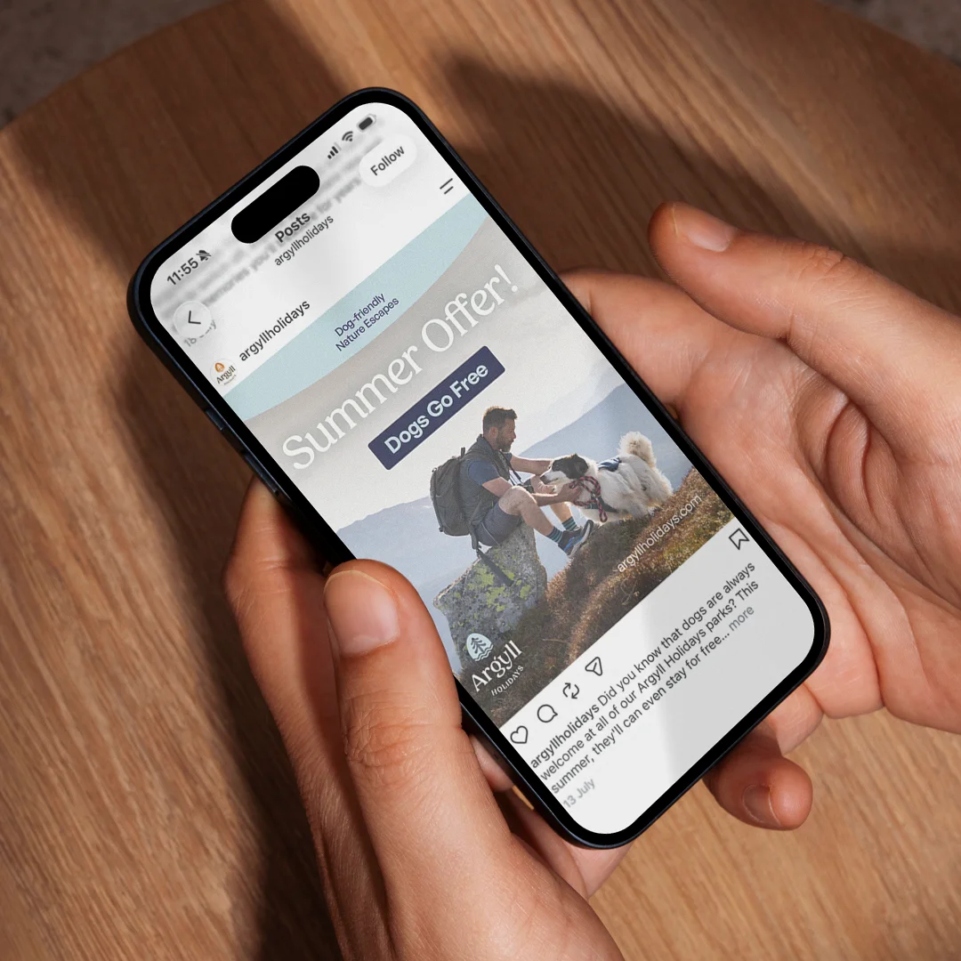 Person holding a smartphone displaying an Instagram post from argyllholidays advertising a summer offer with 'Dogs Go Free', featuring a man sitting on a rock petting a dog in a nature setting with mountains in the background.