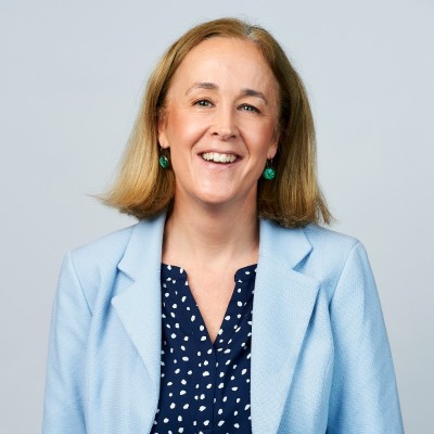 Smiling woman with shoulder-length blonde hair wearing a light blue blazer and dark blue blouse with white dots against gray background.