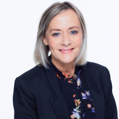 Smiling middle-aged woman with light blonde shoulder-length hair, wearing a dark blazer over a floral blouse, against a plain light background.
