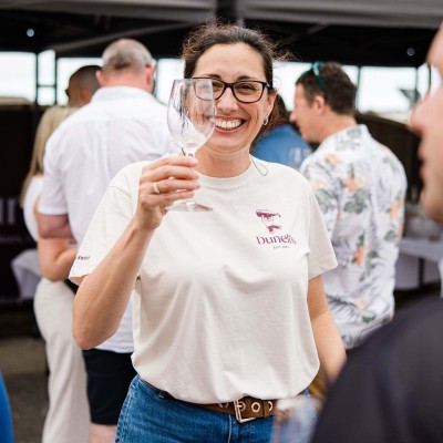 Smiling woman wearing glasses and a white Dunedin shirt holds an empty wine glass at an outdoor social event with people in the background.