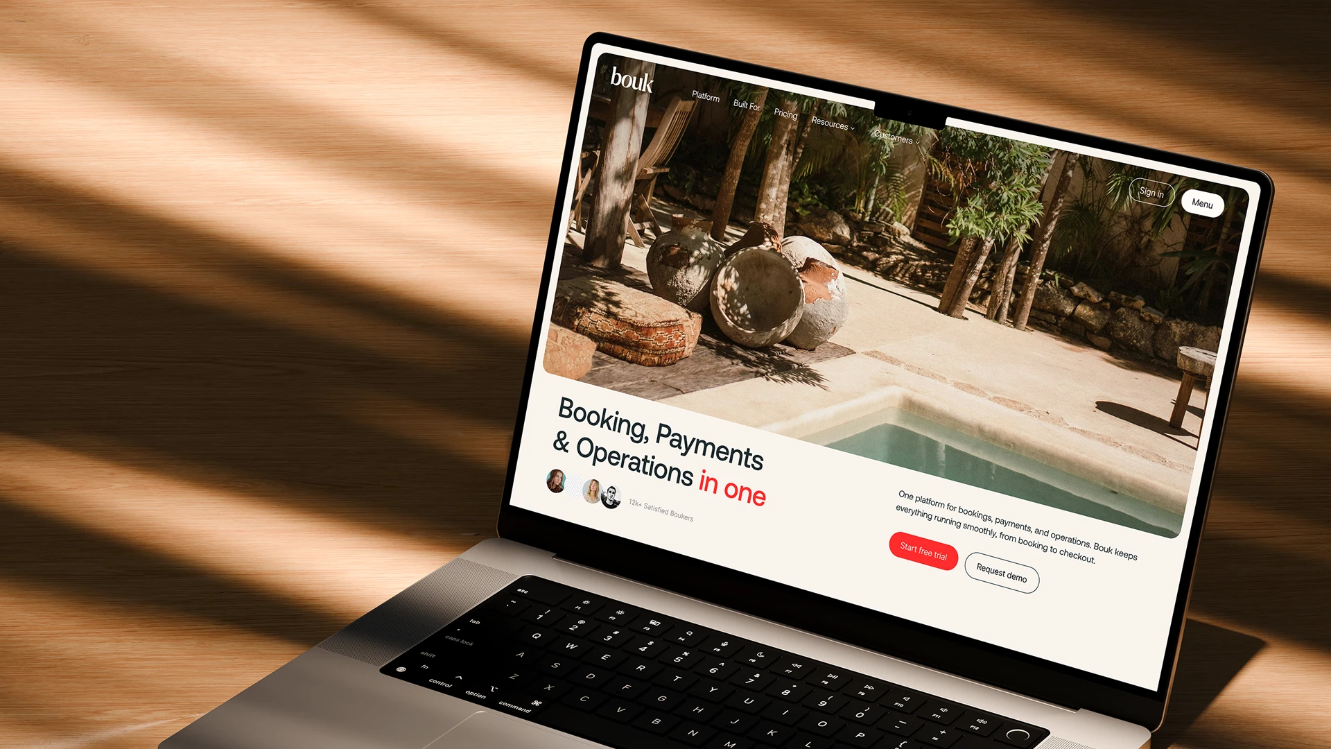 Laptop on wooden table displaying a website homepage for Bouk with text promoting booking, payments, and operations integration, a poolside image, user icons, and buttons offering a free trial and demo request.