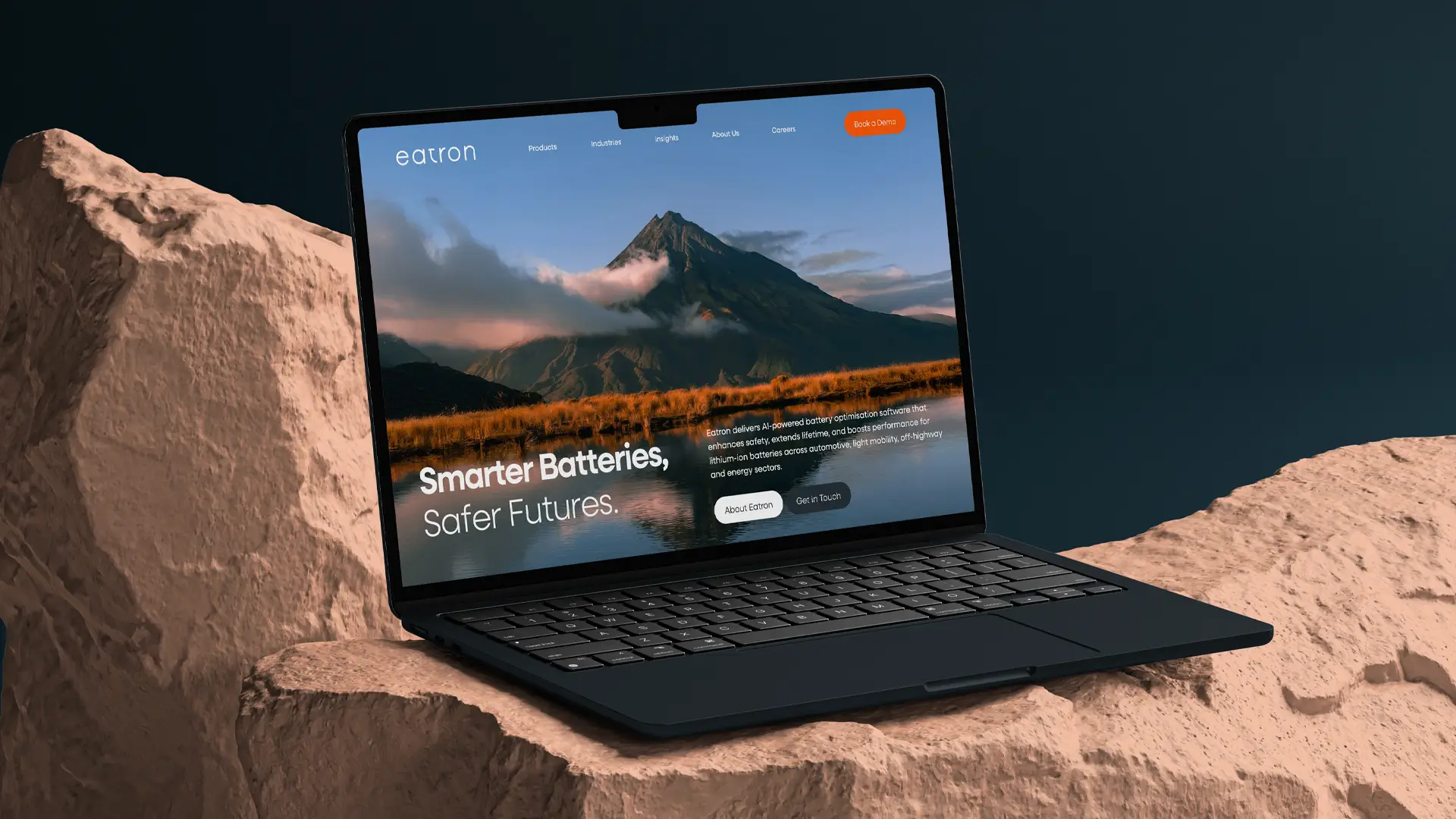 Laptop on textured rock surface displaying Eatron website homepage with a mountain landscape background and text about AI-powered battery optimization.