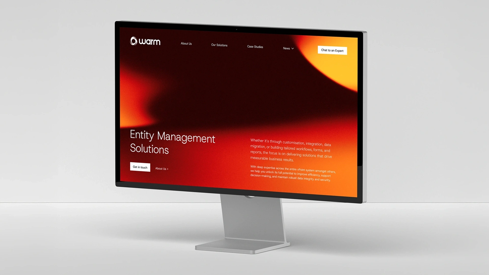 Computer monitor displaying the Warm website homepage with a red and orange gradient background. The page highlights 'Entity Management Solutions' with text describing customization, integration, and data migration services to improve business results and efficiency. Navigation links for About Us, Our Solutions, Case Studies, News, and a Chat to an Expert button are visible.