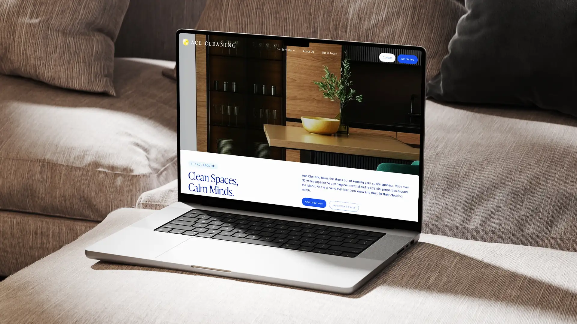 Laptop on a beige couch displaying the Ace Cleaning website homepage with a kitchen background and text promoting clean spaces and calm minds.