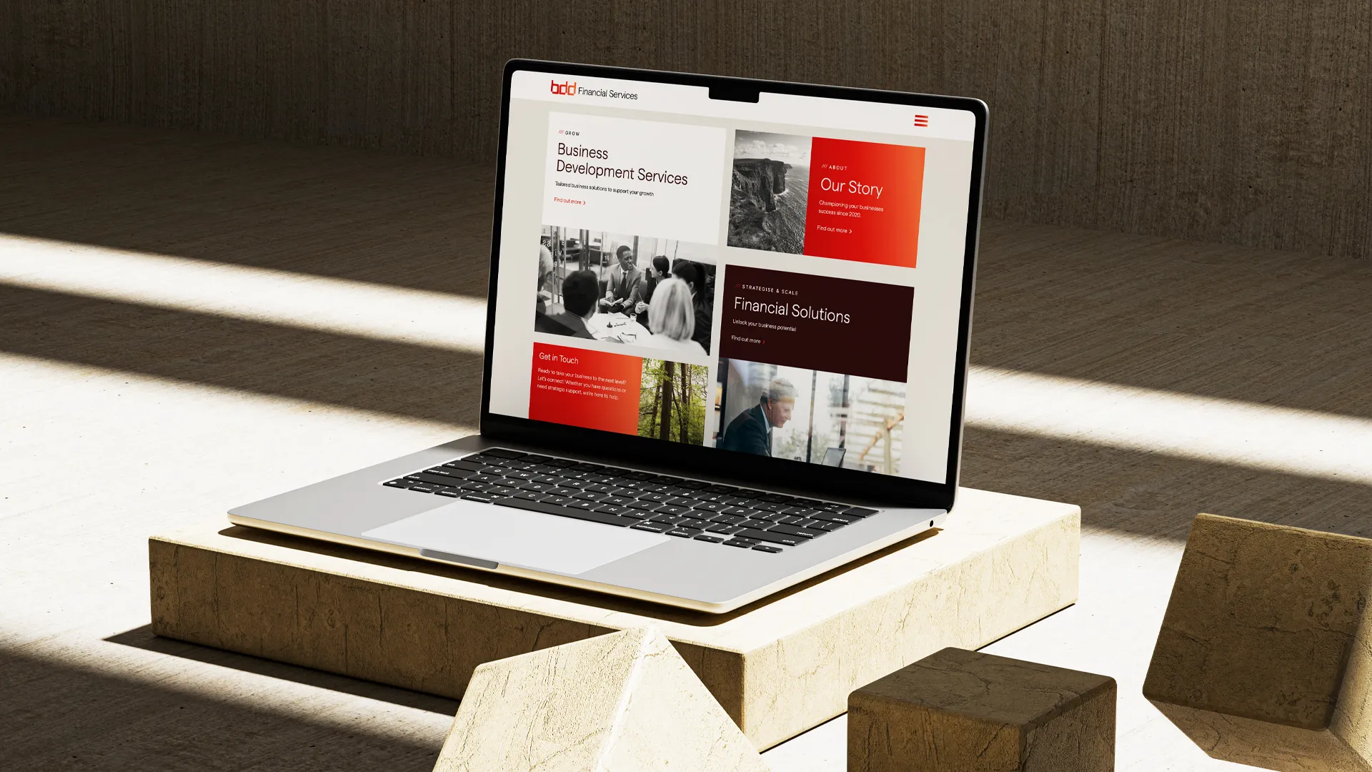 Open laptop on a stone pedestal displaying a business website with sections for Business Development Services, Our Story, Financial Solutions, and Get in Touch, using red, black, and white color blocks.