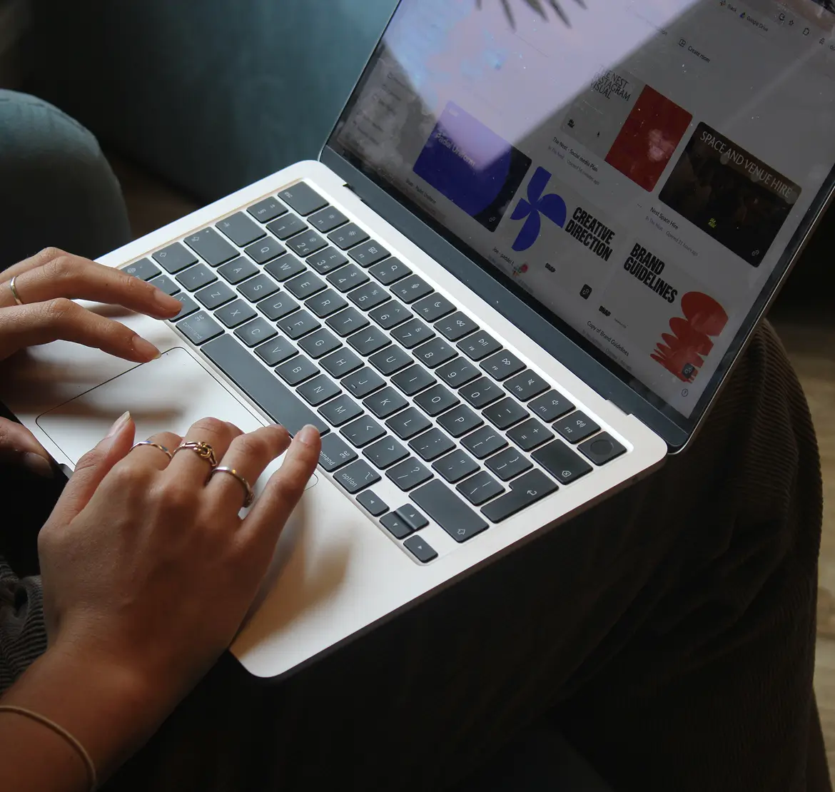 Person typing on a silver laptop with a black keyboard, screen showing design project thumbnails titled 'Creative Direction' and 'Brand Guidelines'.