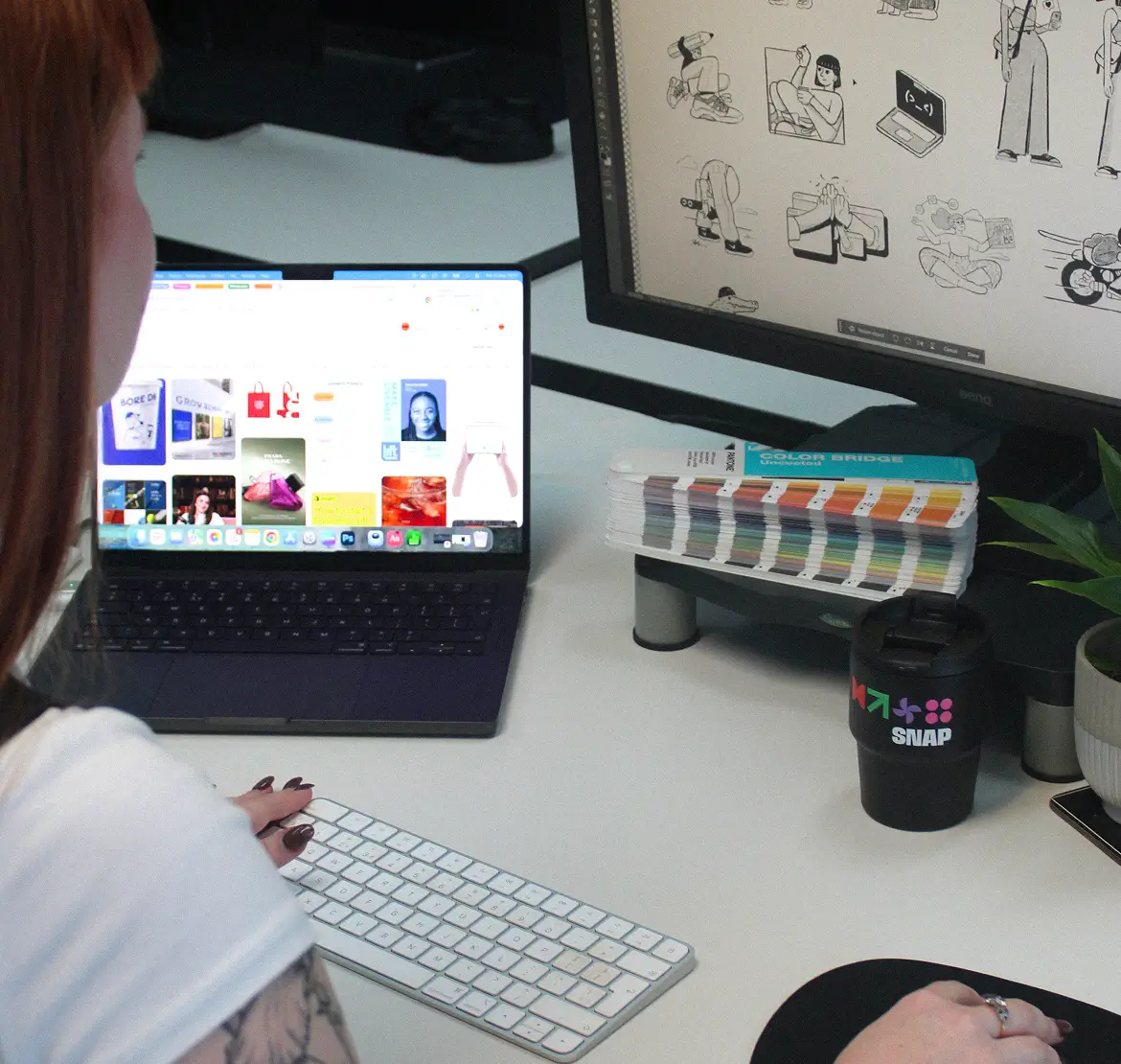 A person with red hair working at a desk using a wireless keyboard and mouse, viewing colorful graphics on a laptop screen and black-and-white illustrations on a larger monitor, with a color swatch book and a black SNAP-branded cup nearby.