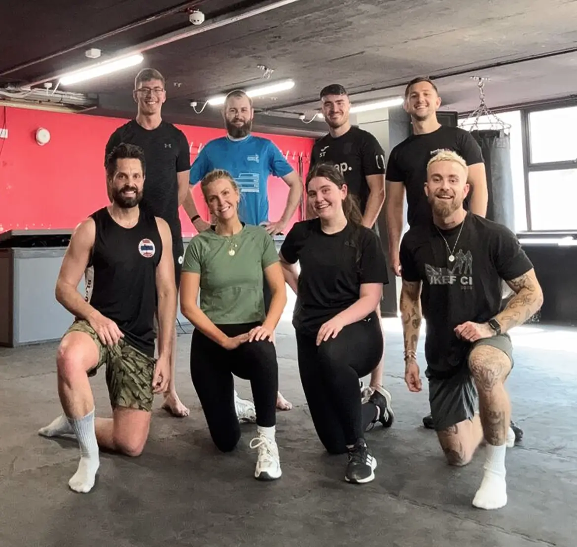 Group of eight adults in casual sportswear posing in a gym, four kneeling in front and four standing behind on a black padded floor with red wall and punching bag in the background.