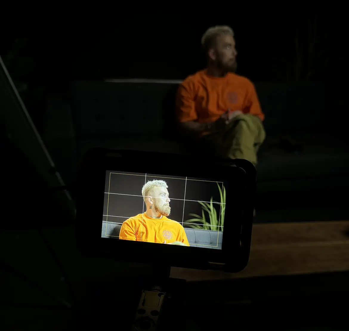 A man with blond hair and beard wearing an orange shirt and light-colored pants is sitting on a dark couch, shown both directly in low light and brightly lit in the display screen of a camera in the foreground.