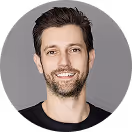 Smiling man with short dark hair and beard wearing a black shirt against a gray background.