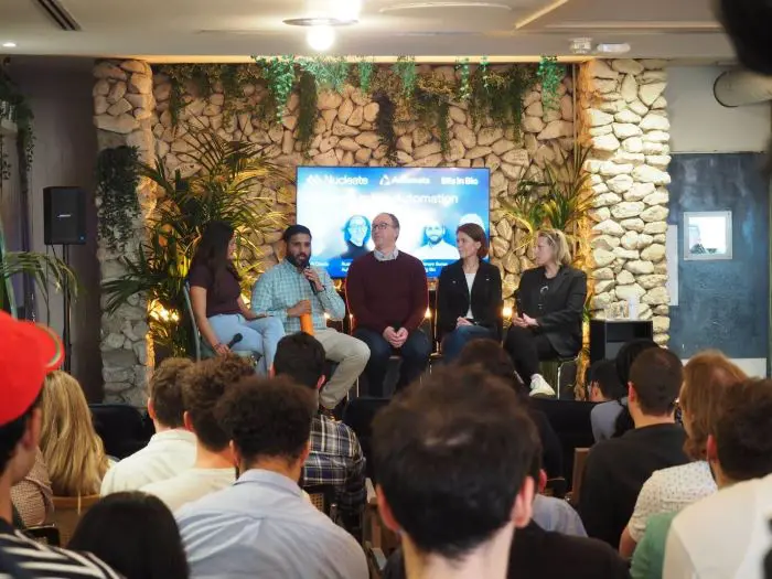 Five people seated on a stage on high stools with a tv screen behind them