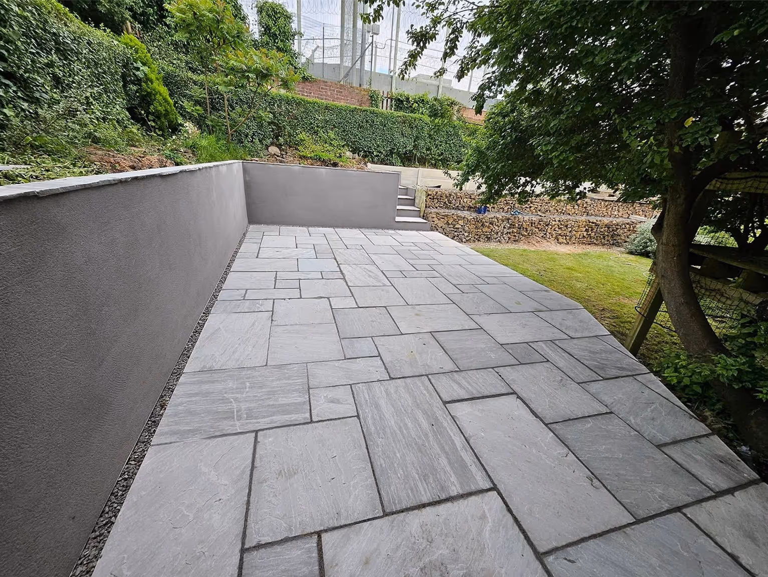 Newly paved gray stone patio with steps leading to a raised garden area surrounded by trees and greenery.