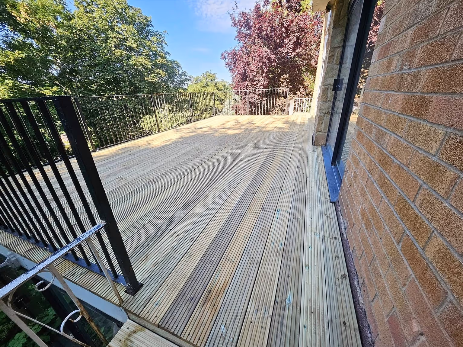 Spacious wooden deck attached to a brick house with black metal railing and surrounded by green trees under a clear blue sky.