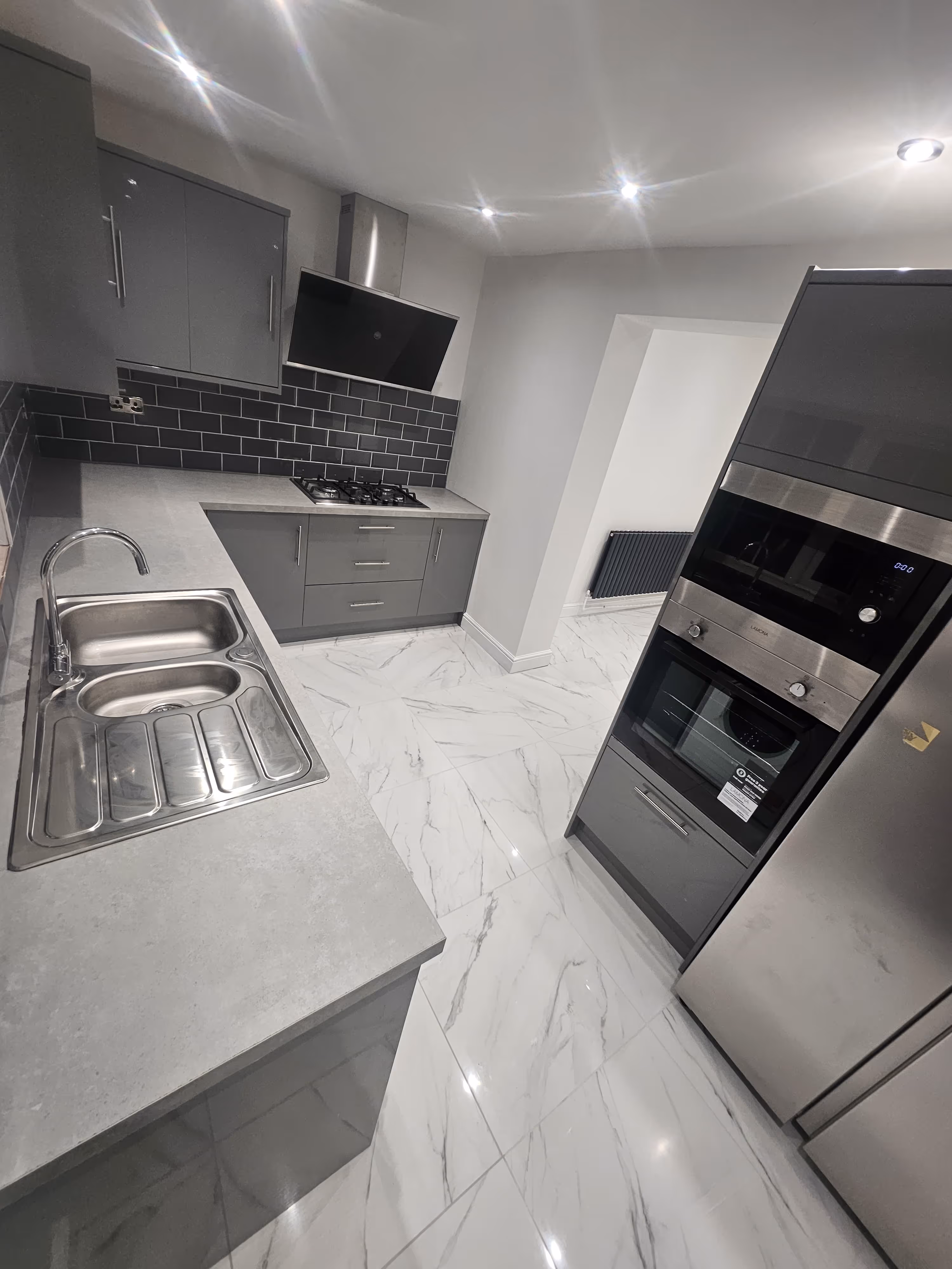 Modern kitchen with grey cabinets, stainless steel sink, stove with black tile backsplash, built-in oven, and marble-patterned floor tiles.