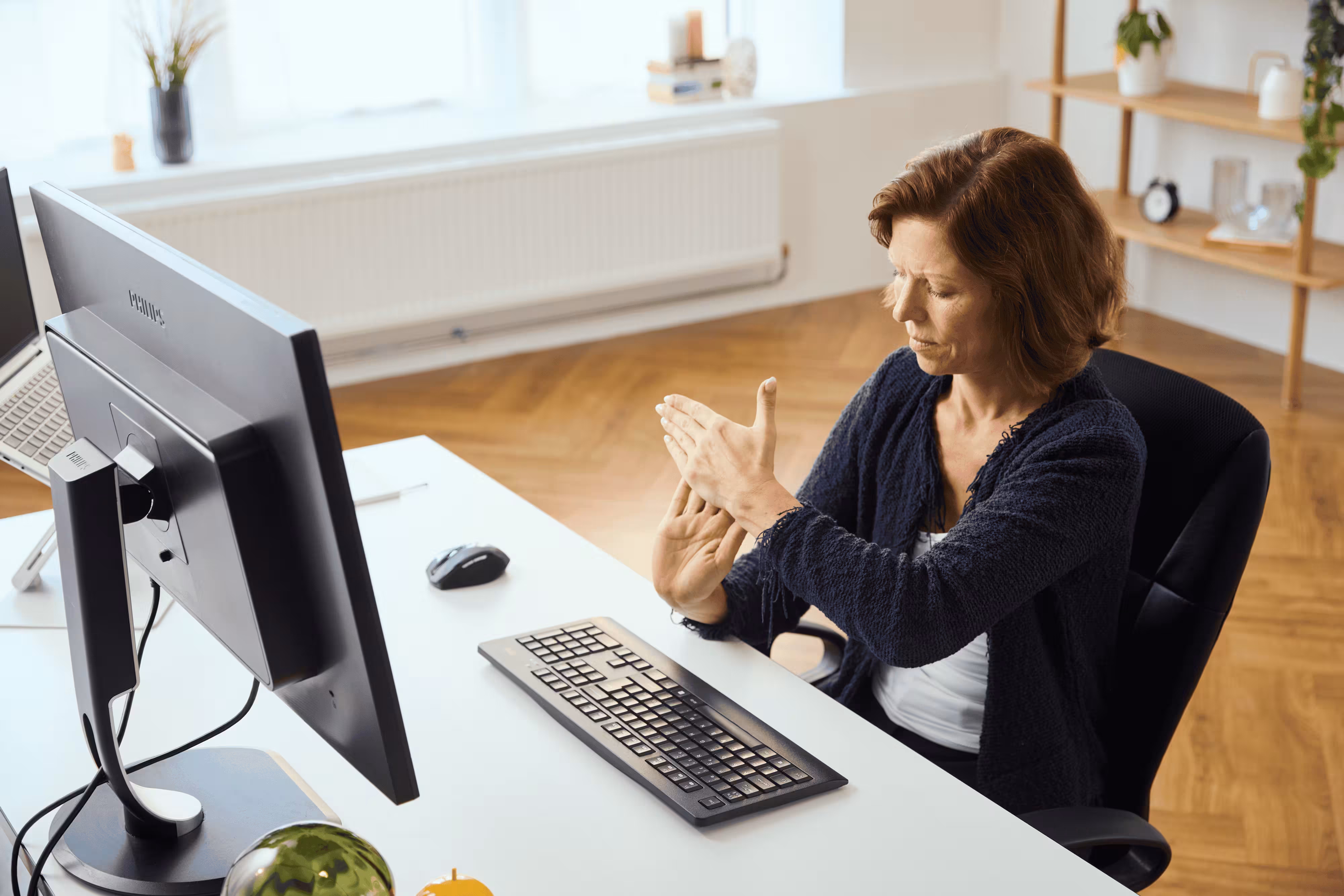 A woman sitting at a desk in front of a computer holds and massages her hand, appearing to experience discomfort. A keyboard and mouse are on the desk, and she is seated in a modern, bright office space.