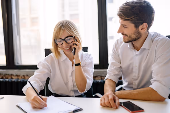 Coworkers at a desk, one writing and one talking on the phone, illustrating everyday office work that can affect hand and wrist comfort