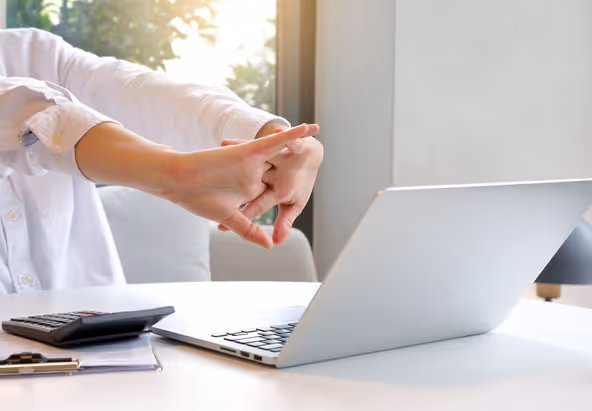 Person stretching their wrist and hand at a laptop, suggesting wrist strain or tendonitis in an office setting