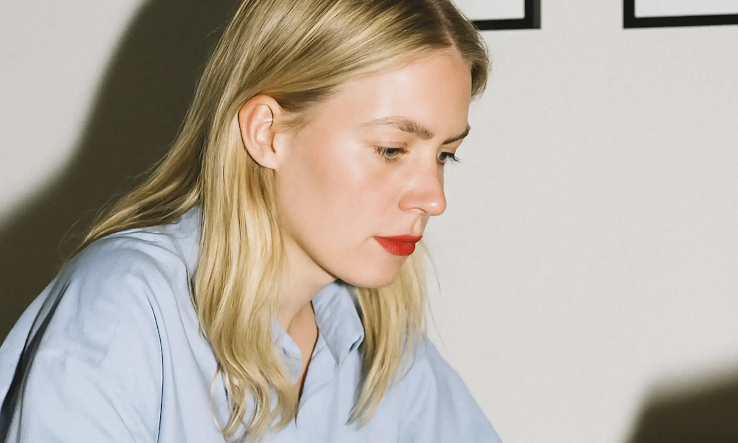 Blonde woman with red lipstick wearing a light blue shirt looking down thoughtfully against a plain background.