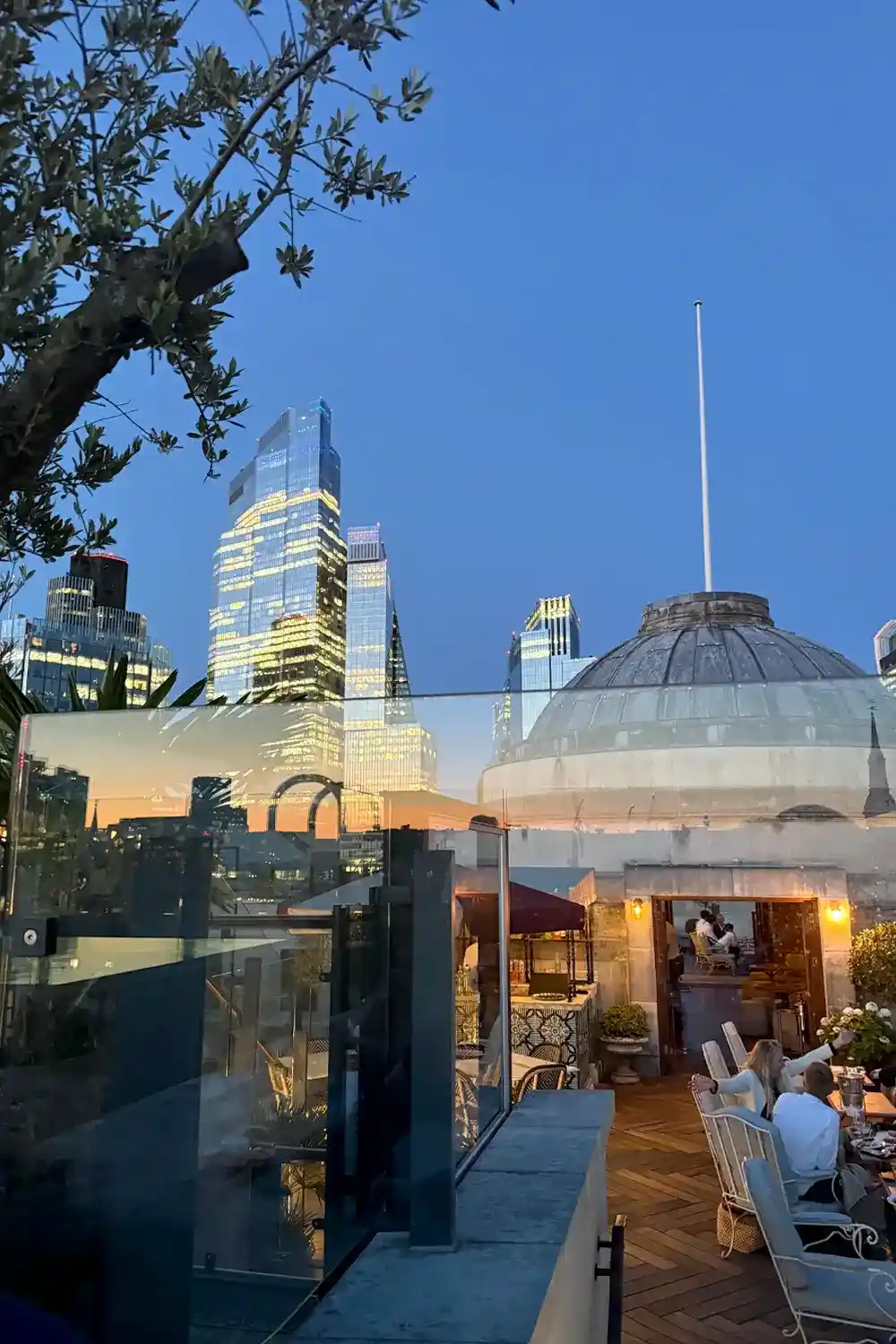 Evening London cityscape viewed from a rooftop terrace with people dining, glass railing, and illuminated skyscrapers reflecting sunset light.