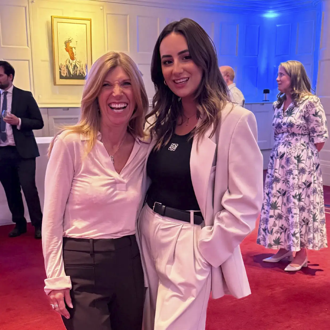 Ellie Hecht and Penny Reason-Goddard smiling and posing together in a well-lit room with red carpet, with other people in the background.