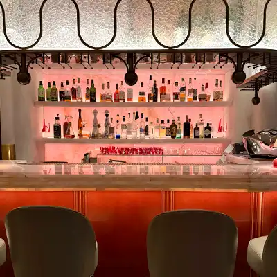 Bar counter with three stools facing shelves of assorted liquor bottles illuminated with pink lighting.