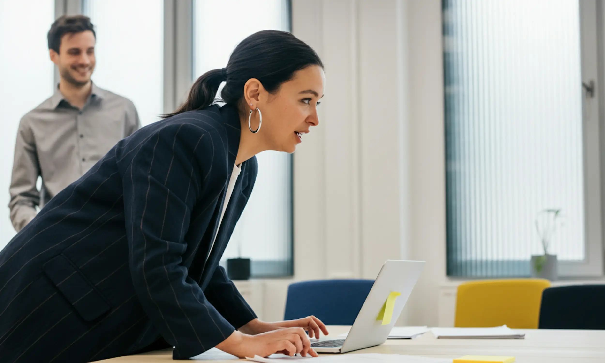 A woman in a blazer leaning over a table and working on a laptop, with a smiling man standing behind her in an office setting.
