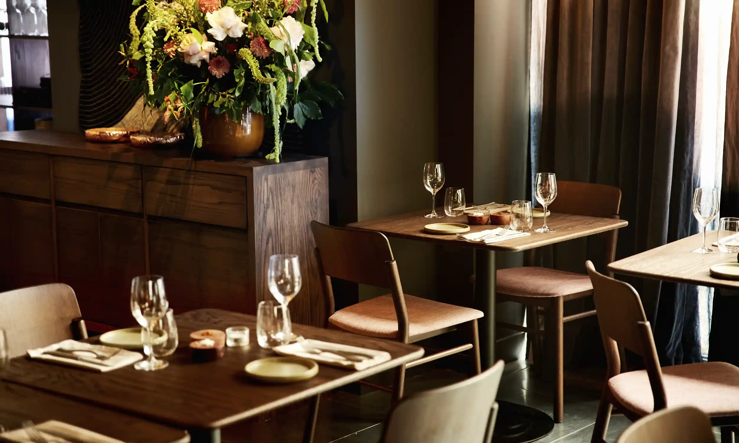 Cozy restaurant interior with wooden tables set with wine glasses, plates, and napkins, next to a cabinet with a large floral arrangement.