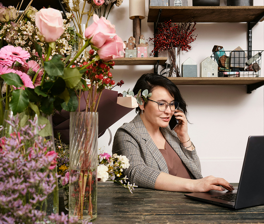 A woman looking at a laptop and talking on a phone in a florist shop.