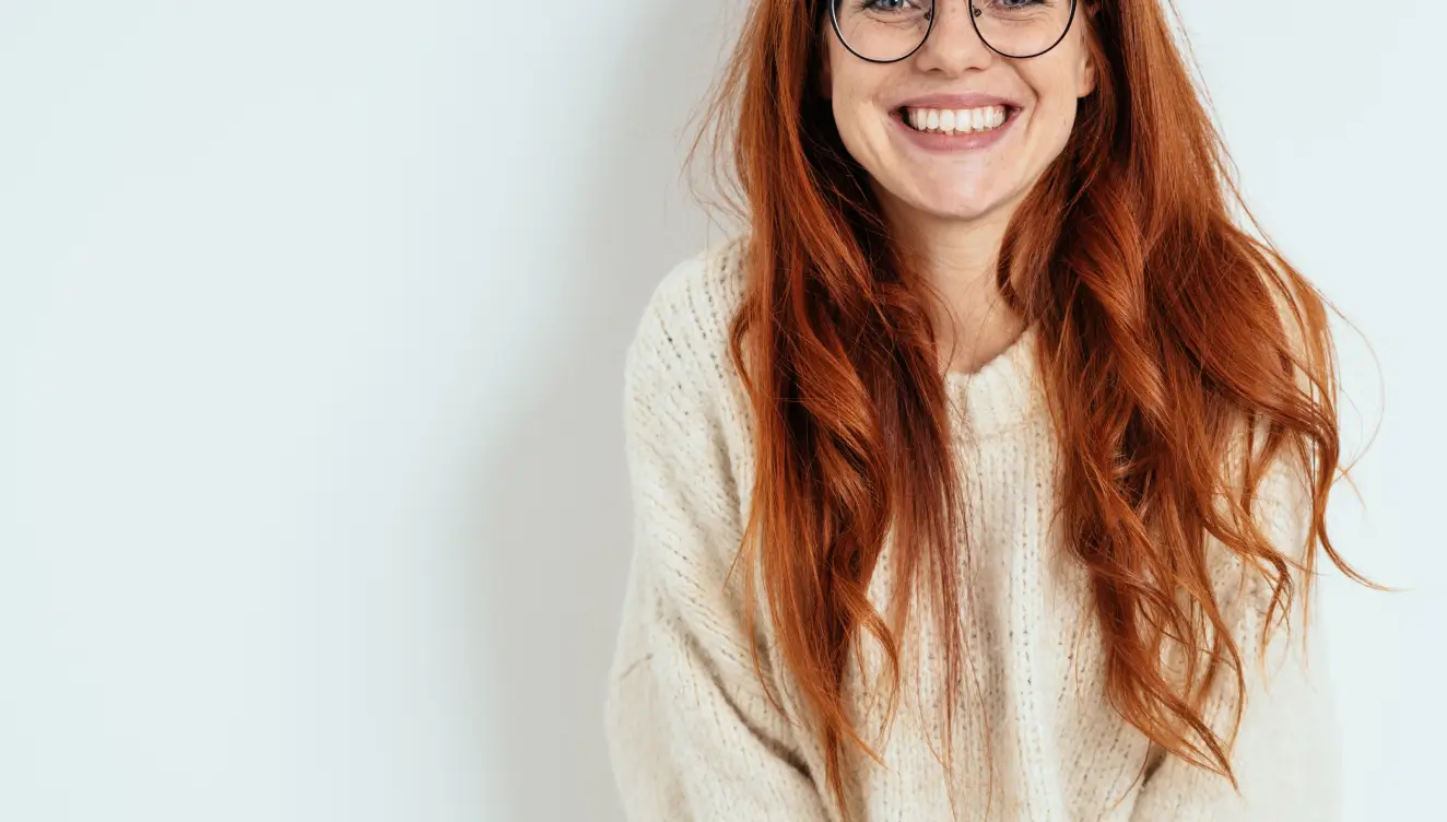 A person with long red hair and glasses smiles while wearing a light sweater.