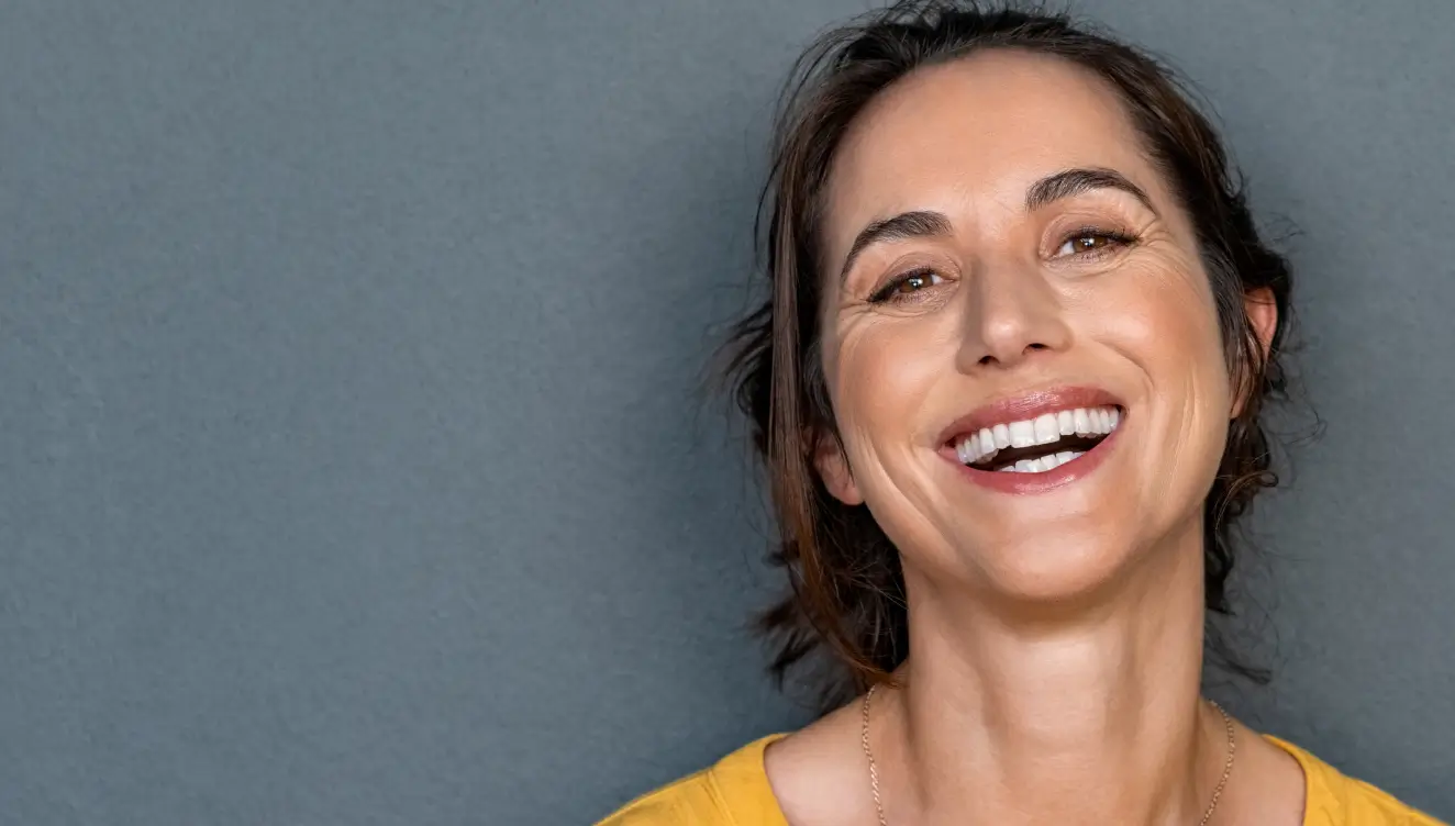 A smiling woman in a yellow shirt stands against a plain gray background.
