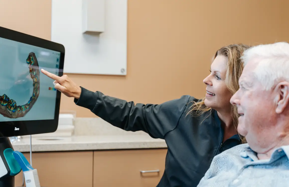 A woman points at a dental image on a screen while explaining to an older man.