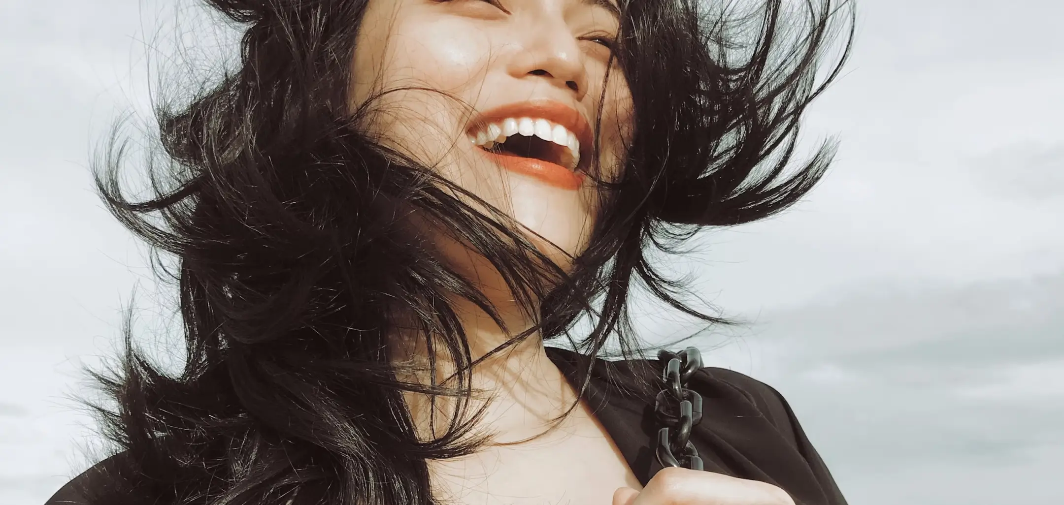 A person with long hair smiles widely, holding a swing chain, wind blowing their hair.