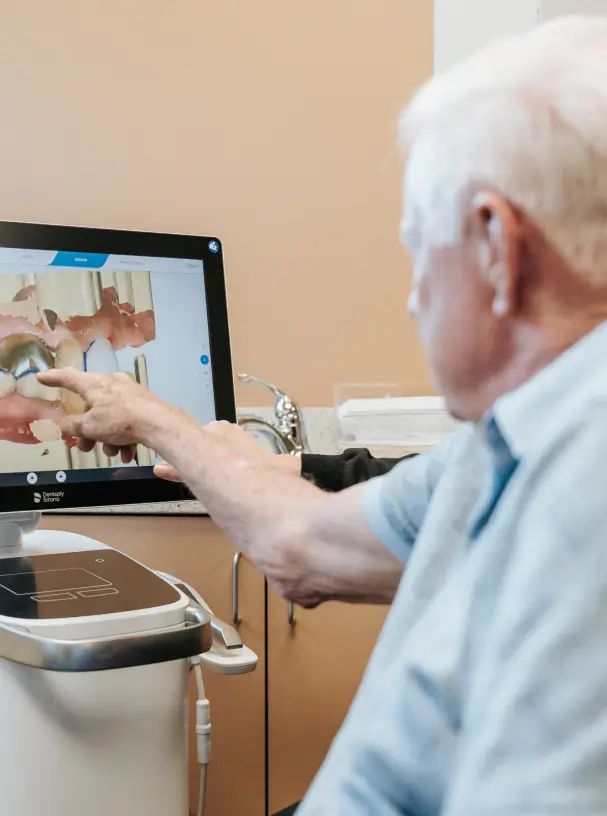 An elderly man points at a dental image on a computer screen in an office setting.