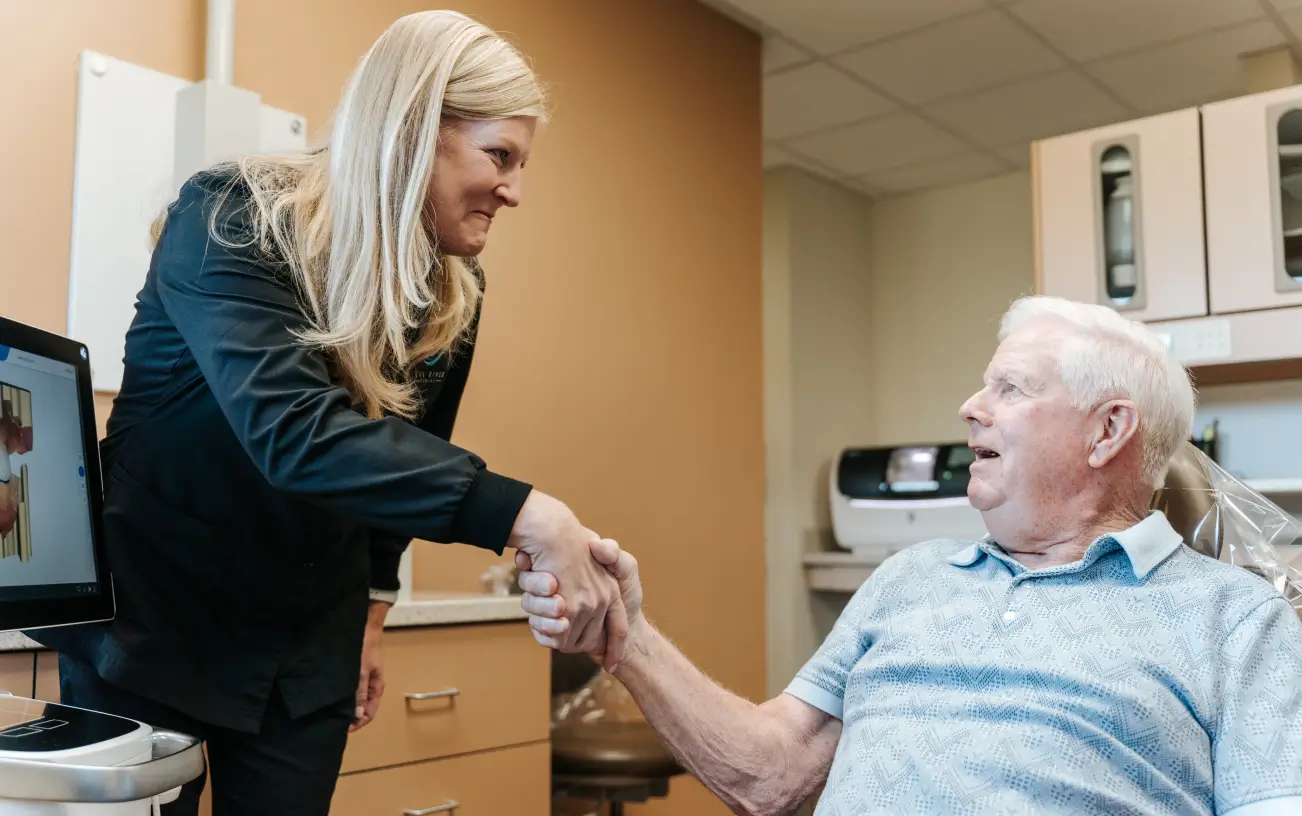 A woman in a black coat shakes hands with an older man sitting in a dental chair.