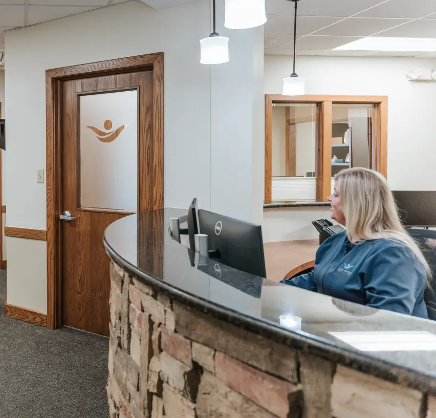 A receptionist sits at a desk with a computer, facing a partially open office door.