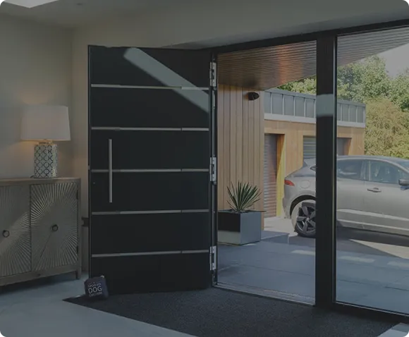Modern black front door open to a driveway with a silver car and wooden garage, interior shows a cabinet with a lamp and a small sign on the floor.
