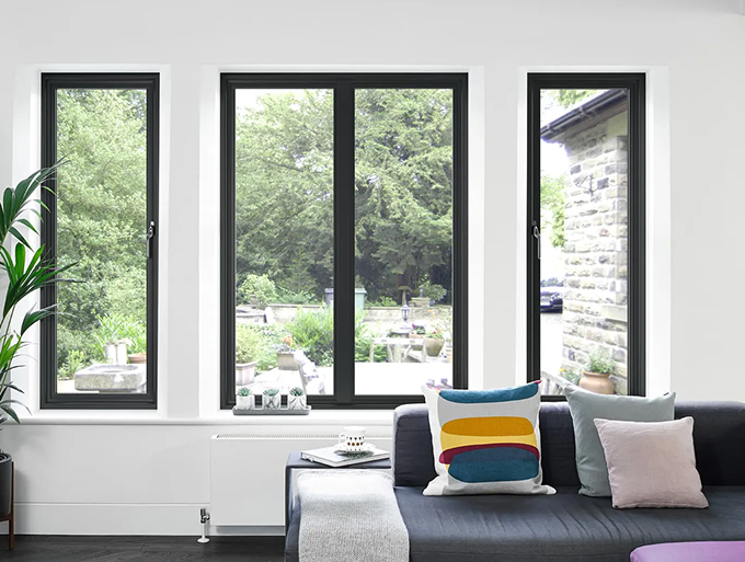 Living room with dark-framed fixed windows overlooking a green garden, featuring a dark sofa with colorful pillows and a white throw.