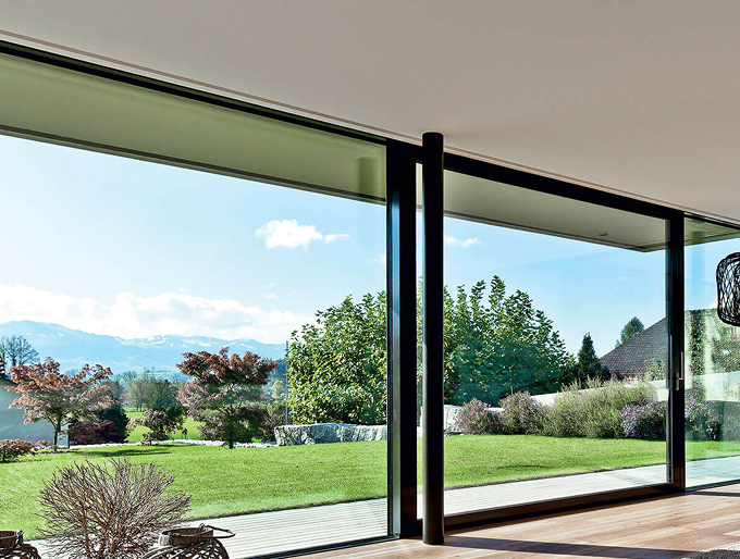View of a garden with green grass, trees, and bushes seen through large sliding glass doors in a modern home.