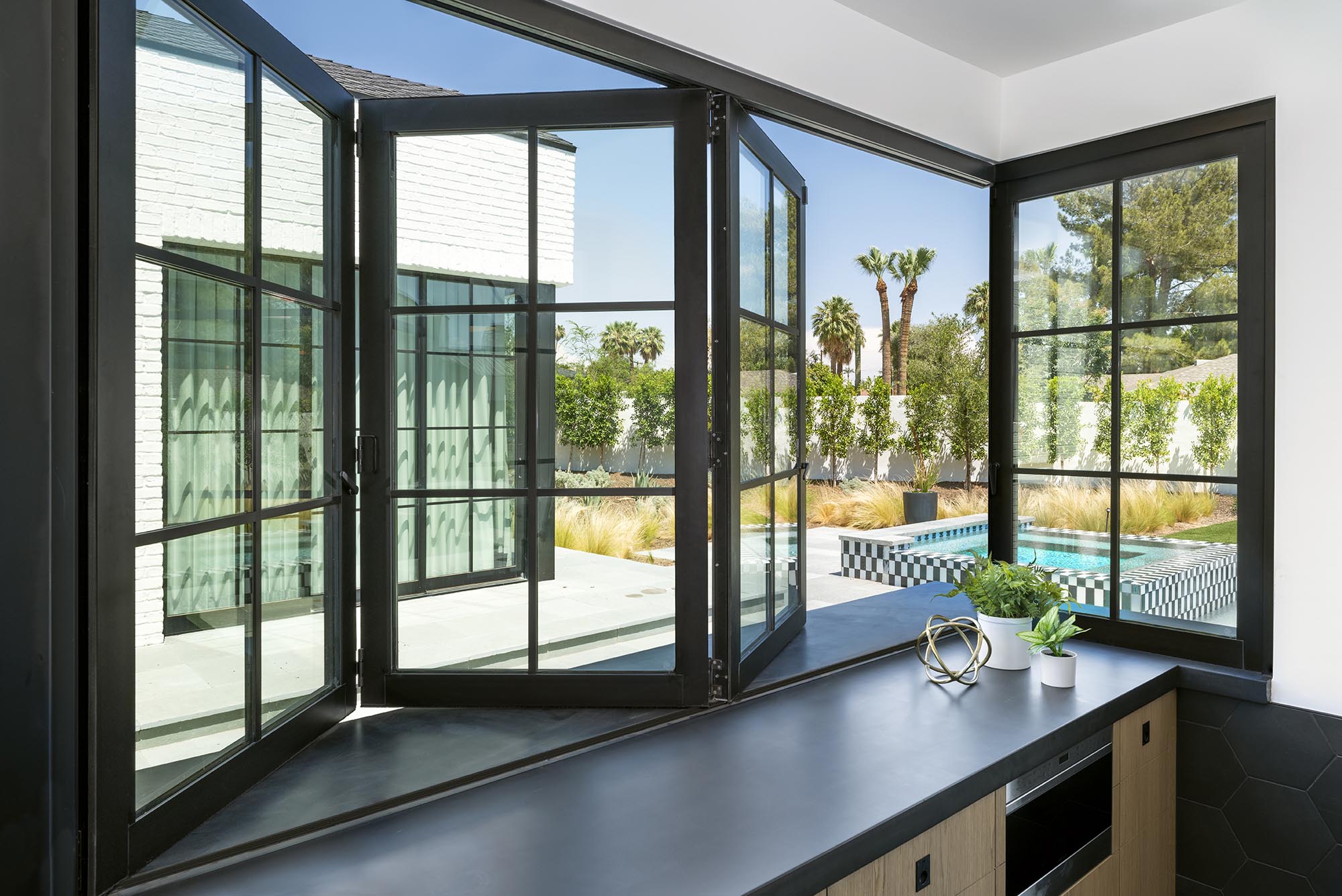 Modern black-framed bi-fold windows opened wide, overlooking a backyard with a tiled pool and palm trees.