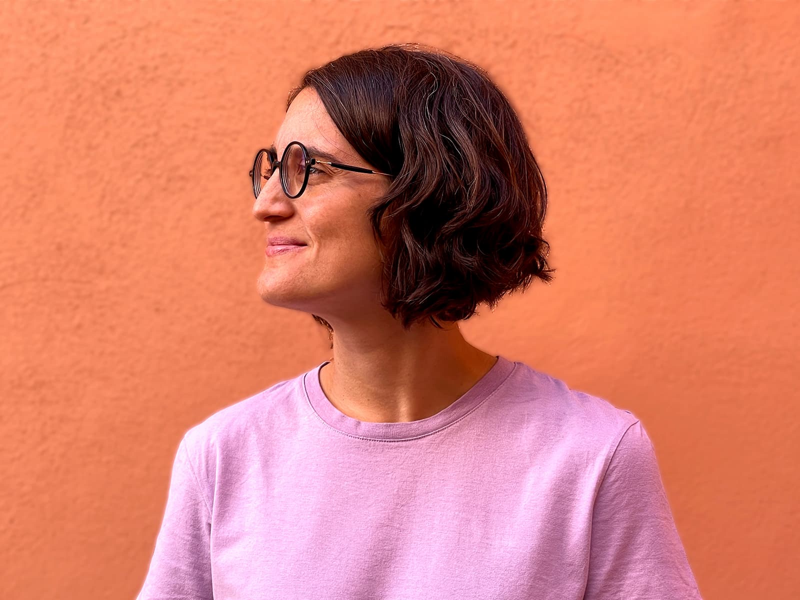 Mujer con gafas redondas y camiseta lila sonríe mirando hacia la izquierda frente a fondo naranja.