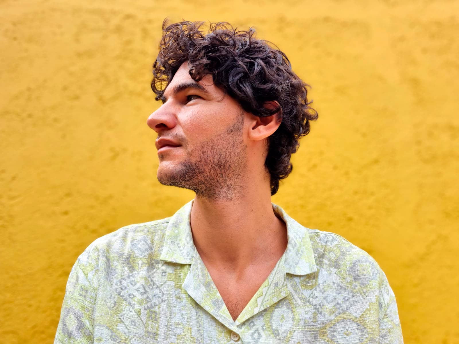 Hombre con cabello rizado y camisa estampada mirando hacia la izquierda con fondo amarillo.