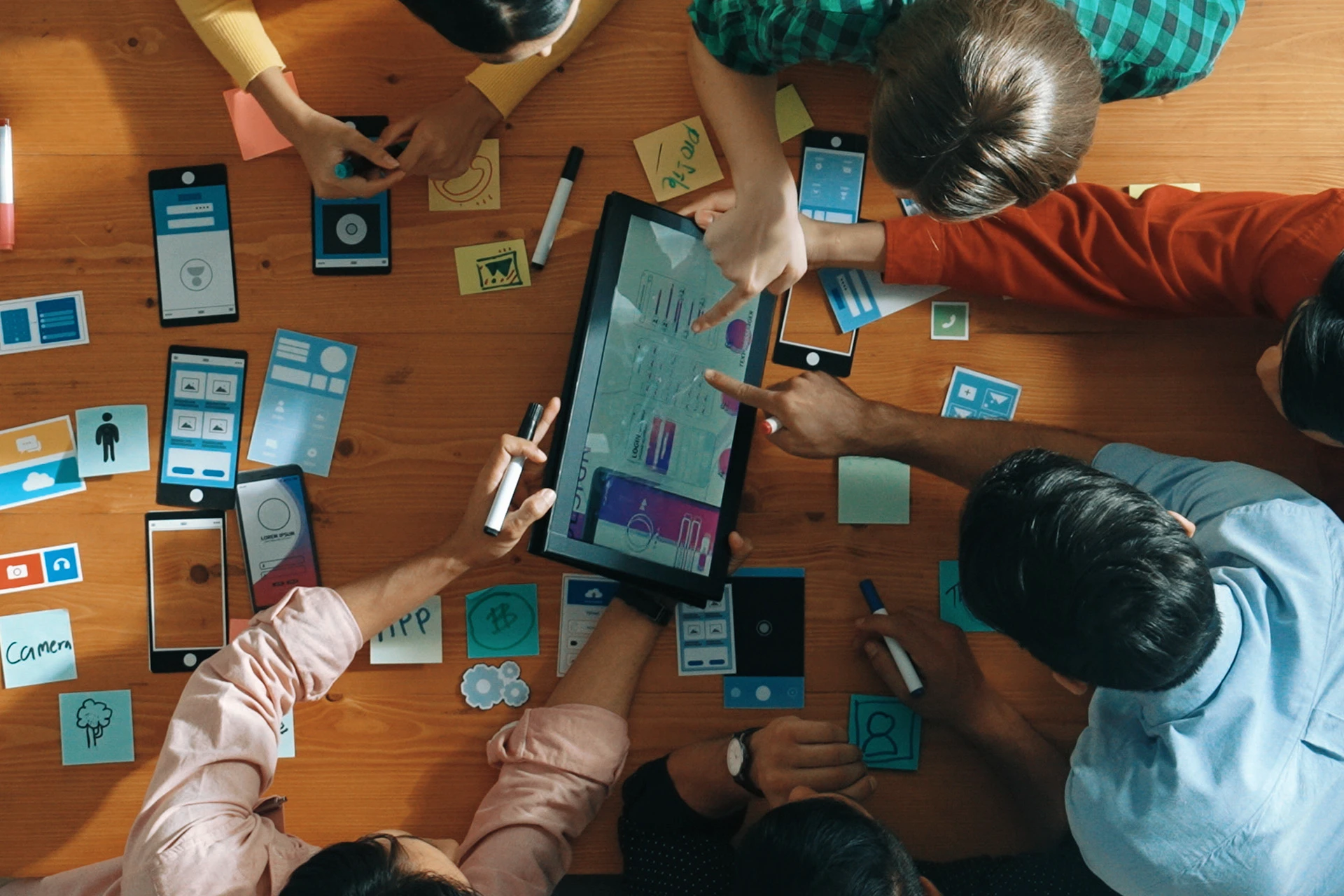 Top view of a group of people around a wooden table collaborating on mobile app design using a tablet and paper prototypes.