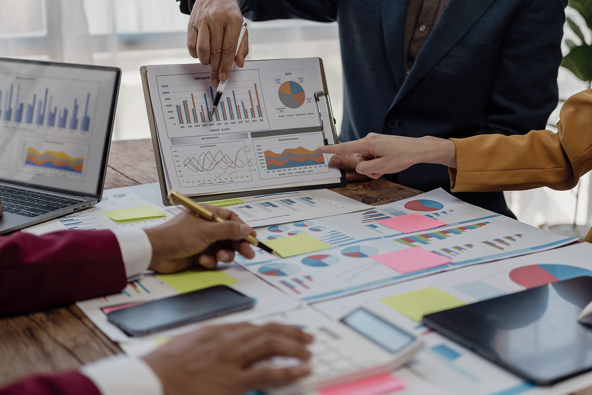Close-up of three people discussing and pointing at charts and graphs on paper and a laptop, analysing business data.