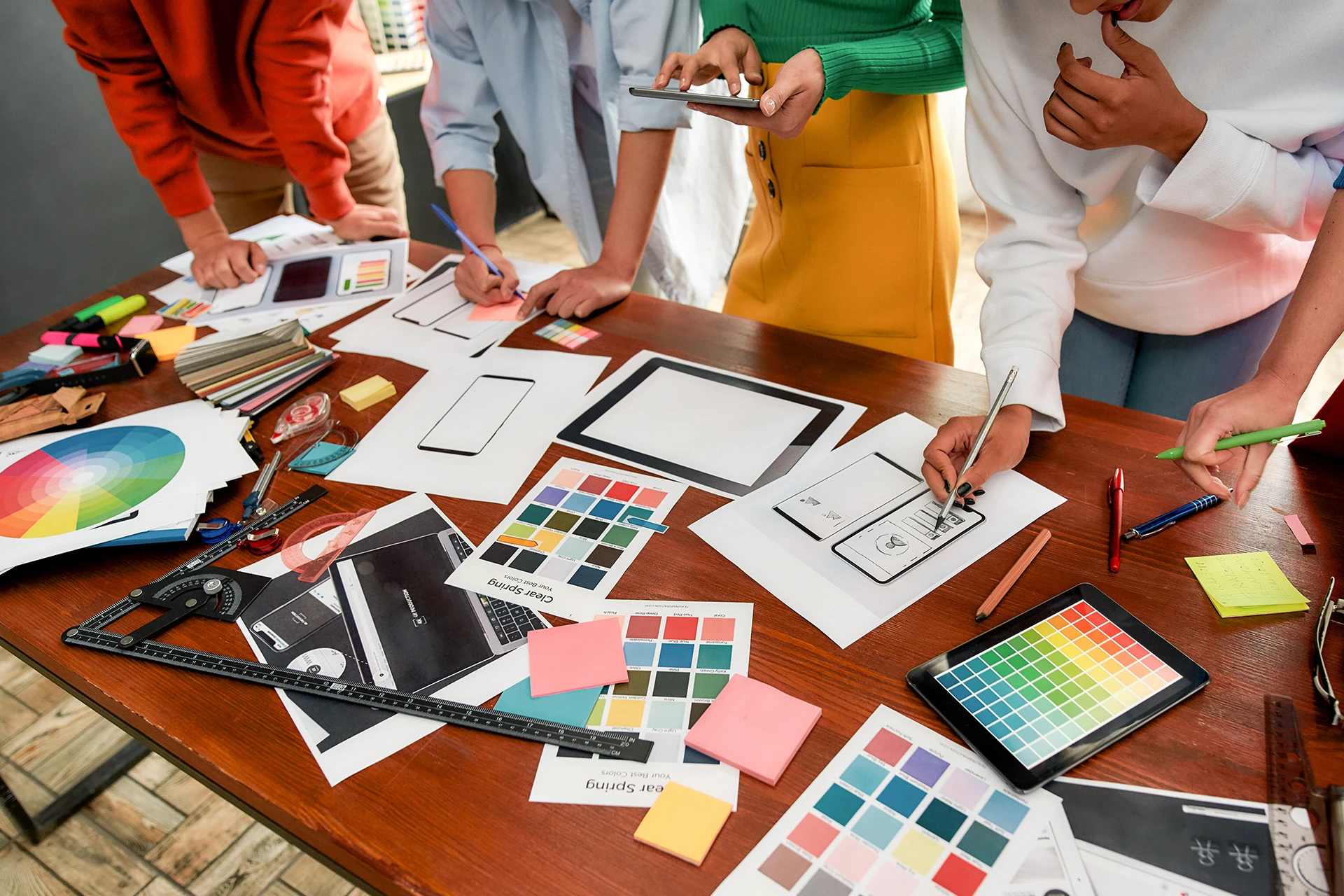 Group of people collaborating on a table filled with colour swatches, design sketches, sticky notes, and digital tablet with colour palette.