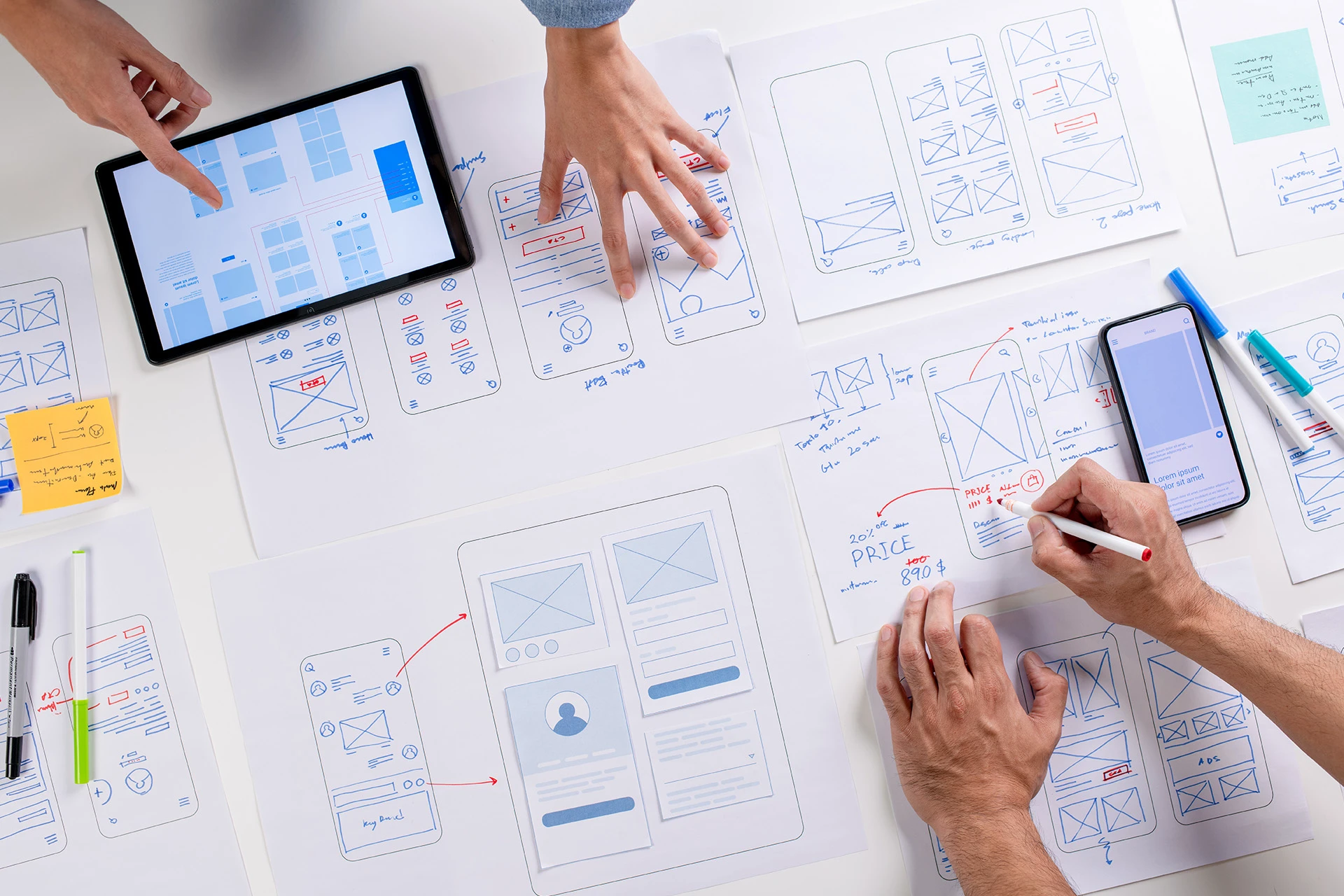 Top view of hands working on mobile app wireframe sketches on paper with tablets and smartphones on a white table.